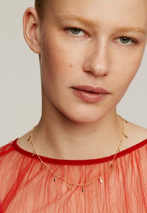Collar de oro con colgante, adornado con acentos en forma de lágrima, llevado sobre una blusa de tela roja transparente con escote redondo y apariencia texturizada.