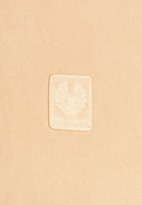 Tessuto beige con una morbida texture, caratterizzato da una patch rettangolare crema embosserata con un emblema alato e il testo "S.C.L. STAFF."
