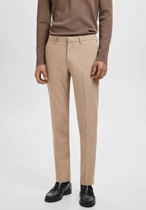 Mann in beigen Anzughosen, schwarzen Lederloafers und einem braunen Langarmshirt, der vor einem schlichten hellen Hintergrund steht.