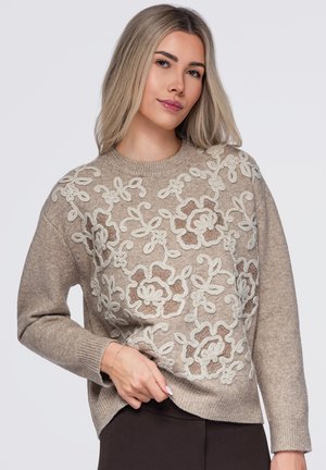 Femme blonde portant un pull beige avec une broderie florale blanche et marron, tenant l'ourlet, sur un fond uni clair.