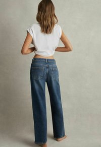 Kvinde med skulderlangt hår står barfodet, iført en hvid cropped top og løstsiddende mørkeblå jeans mod en ensfarvet baggrund.