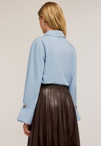 Camicia azzurra a maniche lunghe con colletto, caratterizzata da ampi polsini e bottoni decorativi, abbinata a una gonna plissettata in pelle marrone.