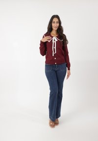 Cárdigan de punto en color burdeos con lazo de cinta blanca, cierre de botones y puños acanalados. Combinado con jeans azules acampanados. Textura suave, diseño informal.