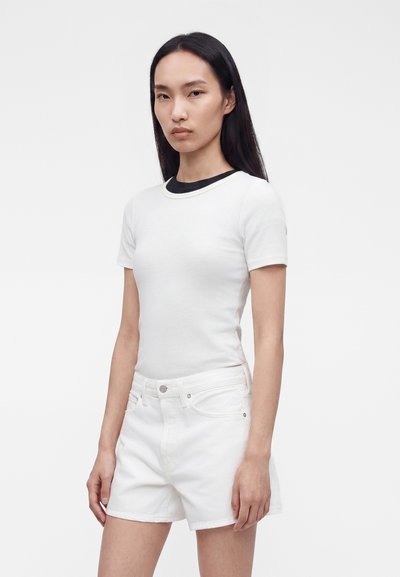 Γυναίκα με μακριά μαύρα μαλλιά που φορά εφαρμοστό λευκό μπλουζάκι πάνω από μαύρο τοπ και λευκά τζιν σορτς, στέκεται μπροστά σε ένα απλό φόντο.