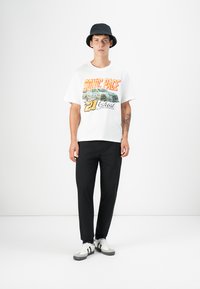 T-shirt graphique blanc avec un texte multicolore et un design de voiture, pantalon noir et chapeau seau noir. Porté avec des baskets blanches et grises.