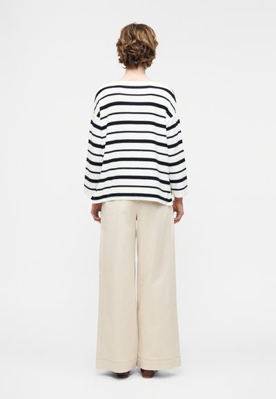 Άτομο με κοντά σγουρά μαλλιά, φοράει λευκό και μαύρο μακρυμάνικο τοπ με οριζόντιες ρίγες και φαρδιά μπεζ παντελόνια, στέκεται με την πλάτη γυρισμένη.