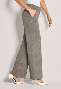 Vrouw in grijze hoge, wijde broek met witte hoge hakken, met �één hand in de zak, staande tegen een neutrale muur.