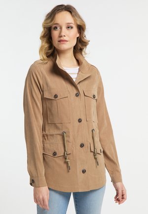 Giovane donna che indossa una giacca beige con bottoni, tasche sul petto e davanti e coulisse in vita, abbinata a jeans azzurri chiari.