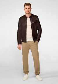 Bruine leren jas met een staande kraag en een ritszak, gecombineerd met een crèmekleurig shirt en kaki chinos, gedragen met witte sneakers.