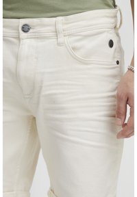 Hvite denimjeans med knappelukking, frontlommer og synlige sømdetaljer. Materialet har en myk, teksturert overflate.