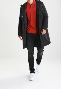 Svart lång kappa, röd polo-skjorta, svarta jeans med slitna detaljer och vita sneakers. Man som går mot en enfärgad bakgrund.