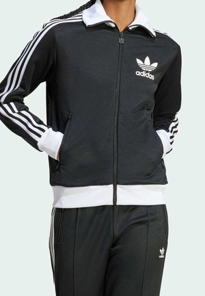 Person trägt schwarzen Adidas-Trainingsanzug mit weißen Streifen an Ärmeln und Hosen, Hände in den Jackentaschen, steht vor neutralem Hintergrund.