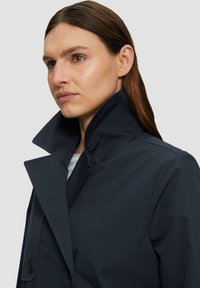 Manteau bleu marine avec un col haut et un design croisé présentant des boutons foncés ; texture lisse, style minimaliste et lignes épurées.