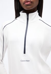 Γυναίκα που φοράει λευκό τοπ Calvin Klein με φερμουάρ στο 1/4, μαύρο περίγραμμα στους ώμους και ψηλό γιακά.