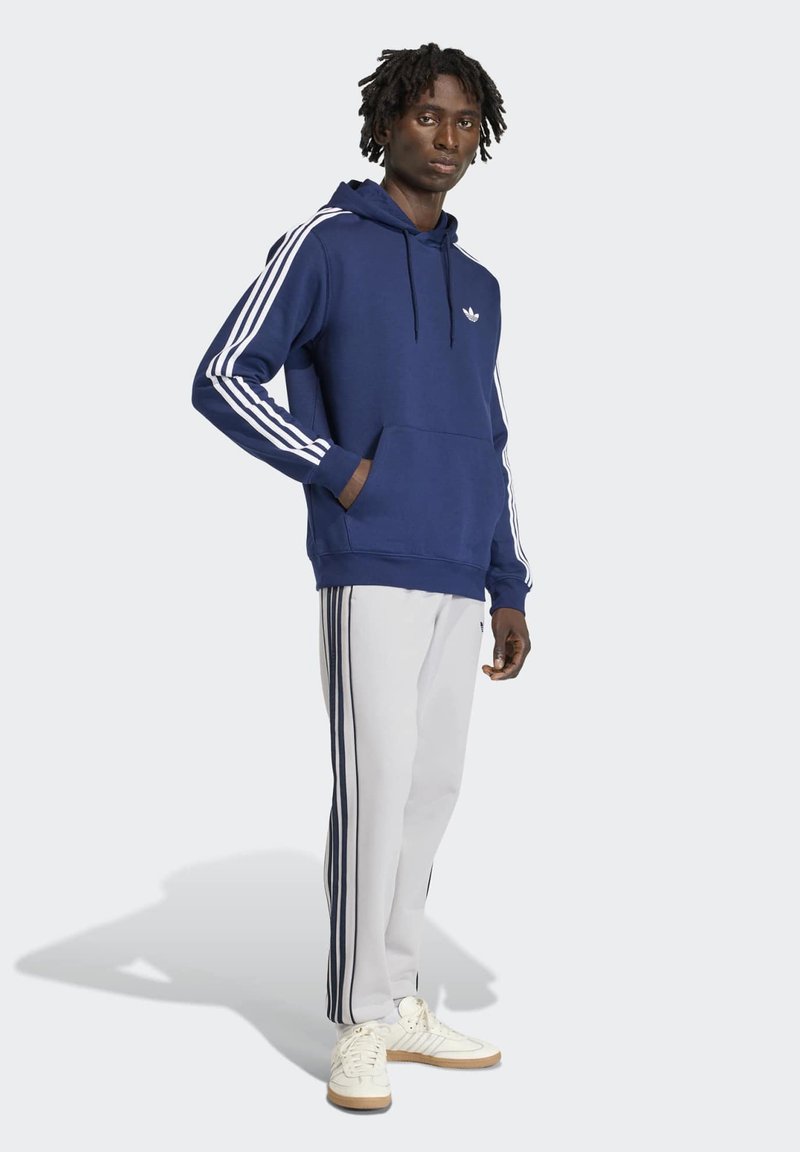 Homme portant un sweat à capuche bleu marine avec des rayures blanches et un pantalon de jogging blanc avec des rayures bleu marine, debout avec une main dans la poche.