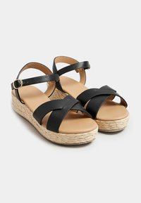 Mustat nahkasaappaat, joissa on juutti-alustat, ristisiteet ja nilkkahihna, sekä vaaleanbeige pohja.