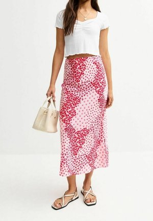 Frau trägt ein weißes Crop-Top, einen rosa-roten Blumen-Midirock, hält eine beige Handtasche und trägt weiße Riemchensandalen.