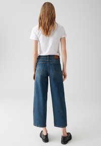 Vrouw met golvend haar, gekleed in een witte shirt met korte mouwen, hoogzekende wijde spijkerbroek en zwarte schoenen, staande tegen een effen achtergrond.