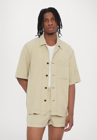 Kurzärmliges, hellkhakifarbenes Hemd mit braunen Knöpfen und einer Brusttasche, kombiniert mit passenden Shorts aus einem glatten Stoff.
