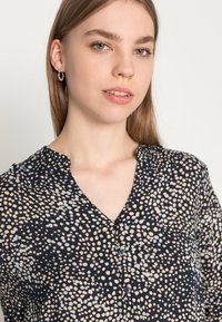 Blusa blu navy con un motivo a pois in crema e azzurro chiaro, caratterizzata da un collo a V, chiusura con bottoni e un discreto dettaglio della tasca sul petto.