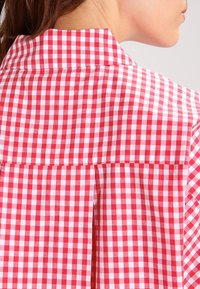 Chemise à col à carreaux rouges et blancs, avec une texture de tissu lisse et un design plissé au dos. Présente un motif vichy évident.