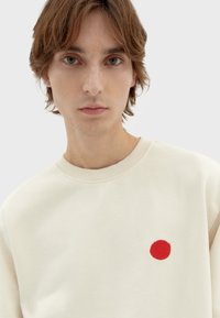 Felpa leggera beige con una toppa rossa ricamata a forma rotonda, caratterizzata da un colletto a costine e una texture morbida. Design semplice e casual.