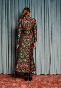 Naine seisab seljaga kaamerasse, kandes rohelist lillelist jopet ja pikka seelikut, tumedad kõrged kontsakingad, taustal sinised kardinad ja pruun vaip.