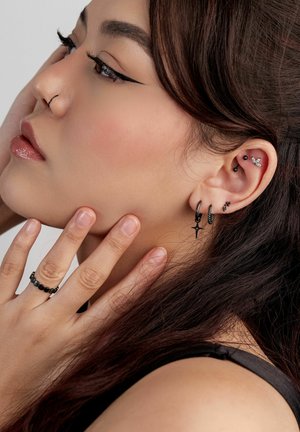 Young woman with dark hair showcasing multiple black and silver ear piercings, a black ring, and winged eyeliner, touching her face gently.