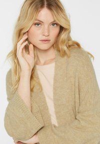 Texturierter beiger Cardigan mit weiten Ärmeln und einem Schalkragen, über einem hellrosafarbenen Top getragen. Weicher Strickstoff, lässiger Schnitt.