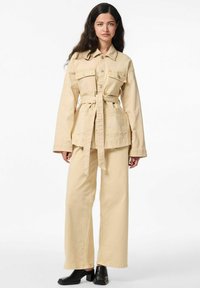 Veste en denim beige avec poches poitrine et ceinture, associée à un pantalon large assorti. Le mannequin porte des chaussures à talons carrés noires.