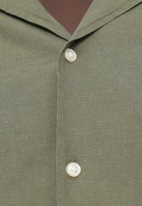 Πουκάμισο σε ελιά πράσινο χρώμα με γιακά σε σχήμα V, υφασμα με υφή και δύο λευκά κουμπιά κοντά στον γιακά, με καθαρές ραφές.