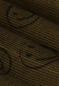 Bruine ribstof met verticale ribbels, bedrukt met kleine smiley gezichten in het zwart. De textuur lijkt zacht en gestructureerd.