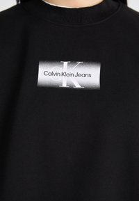 Svart sweatshirt med rund halsringning som har en rektangulär vit etikett. Etiketten visar "Calvin Klein Jeans" i svart och grå text.