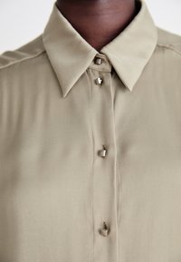 Olivenfarget bluse med krage, knappestolpe med metallknapper, og glatt stoffstruktur.