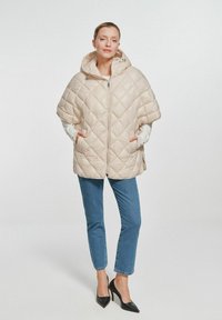Beżowa puchowa kurtka z pikowaniem, z kapturem, zapinana na zamek błyskawiczny, z bocznymi kieszeniami. Noszona na białą bluzkę, w zestawie z niebieskimi dżinsami i czarnymi szpilkami.