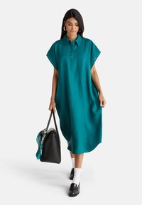 Miętowa sukienka koszulowa z kołnierzykiem, krótkimi rękawami i zapięciem na guziki. Charakteryzuje się luźnym krojem i bocznymi kieszeniami; zestawiona z czarną torebką.