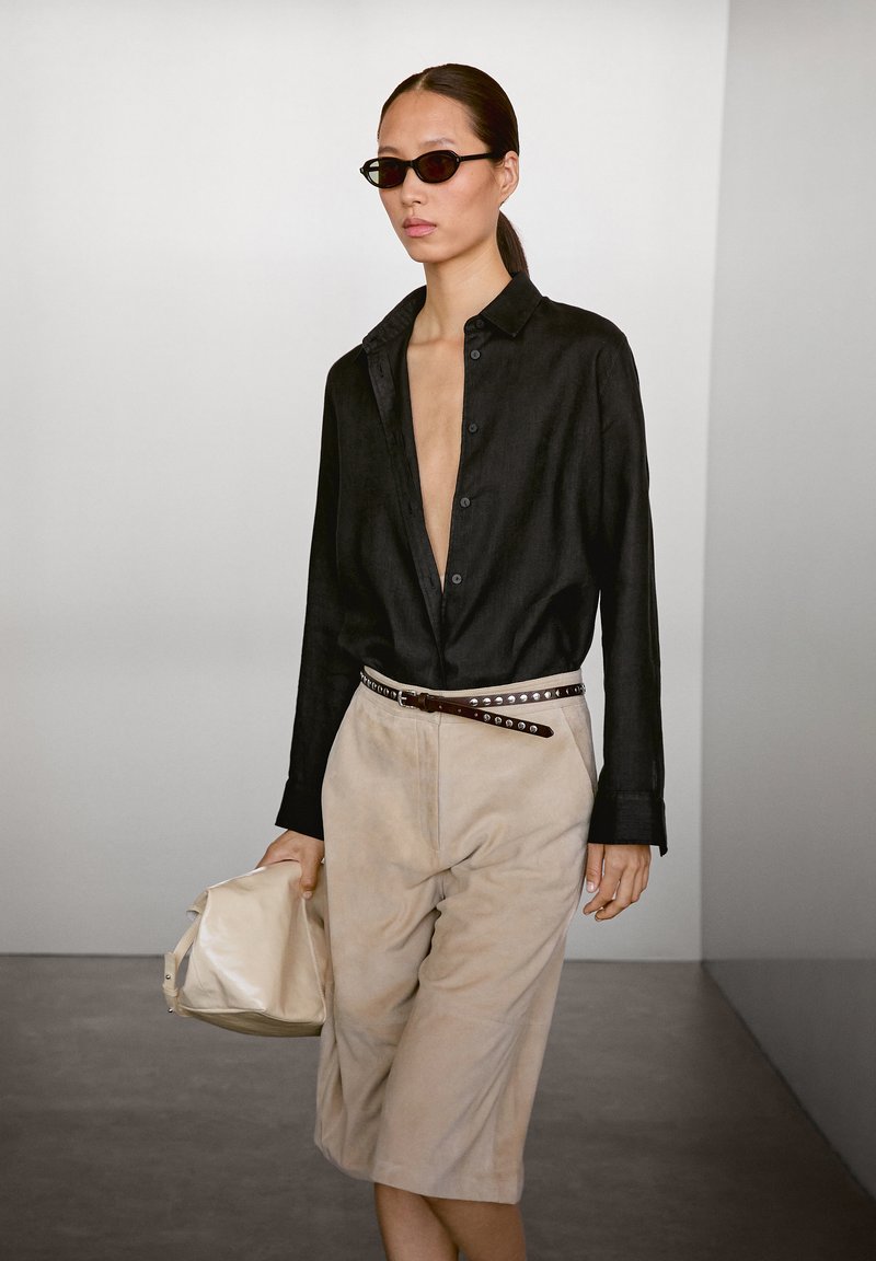 Schwarzes Leinenhemd mit tiefem Ausschnitt, beige Wildledershorts und einem braunen Ledergürtel mit silbernen Nieten. Trägt eine beiges Tasche.