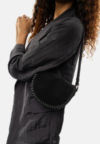 Zwarte suède halve maan schoudertas met geweven randen en een gestructureerde afwerking, gedragen met een donkere, licht gekreukte lange mouwen blouse.