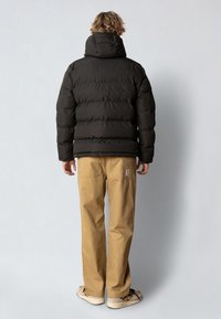 Giacca a vento nera con cappuccio, caratterizzata da un design trapuntato. Abbinata a pantaloni marrone chiaro e scarpe casual. La texture appare liscia e voluminoso.