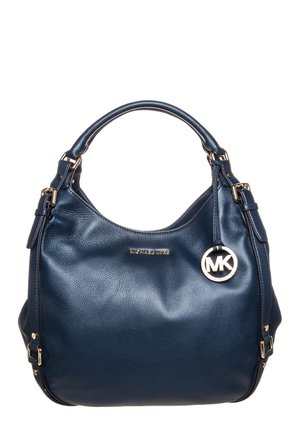 Sac à main en cuir bleu foncé avec une anse unique, une quincaillerie dorée et un pendentif rond avec le logo Michael Kors suspendu sur le côté.