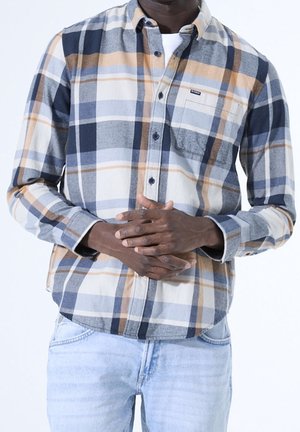 Mand iført en blå, beige og hvid ternet skjorte med knapper og lyseblå jeans, hænderne foldet foran ved taljen.