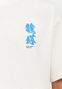 White short-sleeve t-shirt featuring blue Asian-inspired text and "SOUTH TOKYO HARAJUKU" in black, crafted from soft cotton fabric.