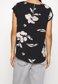 Blusa nera con maniche corte, caratterizzata da ampi motivi floreali bianchi e piccoli accenti rosa, chiusura con zip sul retro, tessuto morbido.