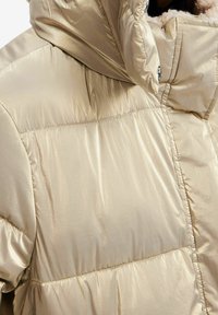 Vadderad, beige puffjacka med en glänsande, metallisk finish. Har en hög krage och synlig sömnad på tygpanelerna.