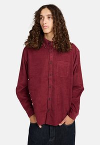 Burgunderfarbendes Cordshirt mit Button-Down-Kragen, Brusttasche und langen Ärmeln, kombiniert mit dunklen Jeans.