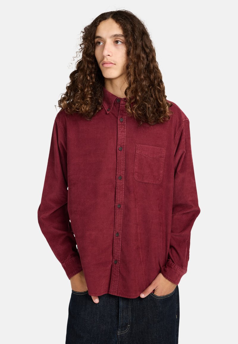 Burgunderfarbendes Cordshirt mit Button-Down-Kragen, Brusttasche und langen Ärmeln, kombiniert mit dunklen Jeans.