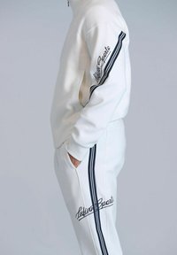 Weißes Trainingsanzug-Set mit hohem Kragen, featuring schwarze Streifenakzente an den Ärmeln und der Hose. Besticktes Logo an der Seite.