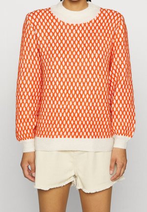 Personne portant un pull orange et blanc à motifs avec poignets et ourlet côtelés, associé à un short en jean écru effiloché.