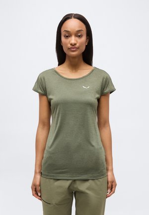 Žena stojící čelem vpřed, oblečená v krátkosleevové olivově zelené košili a kalhotách stejné barvy na jednoduchém světlem pozadí.