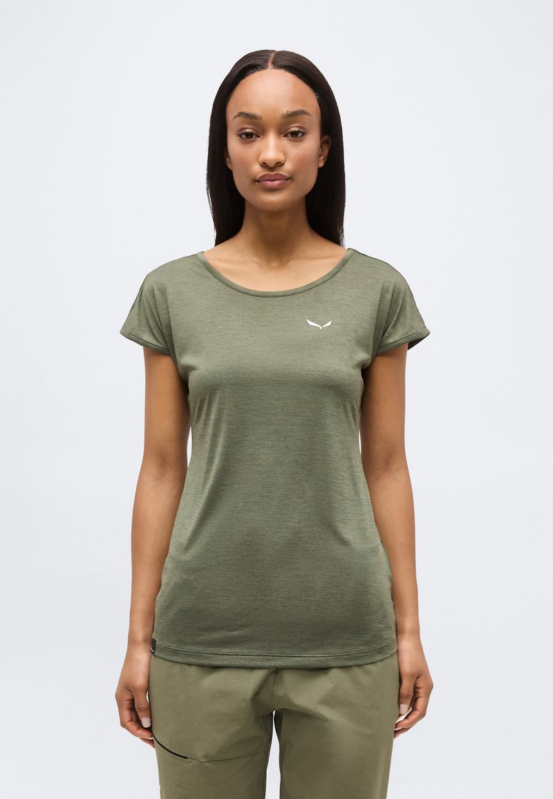 Γυναίκα που στέκεται στραμμένη προς τα εμπρός, φοράει πουκάμισο κοντομάνικο σε ελιά πράσινο και παντελόνι που ταιριάζει, πάνω σε ένα απλό φωτεινό φόντο.